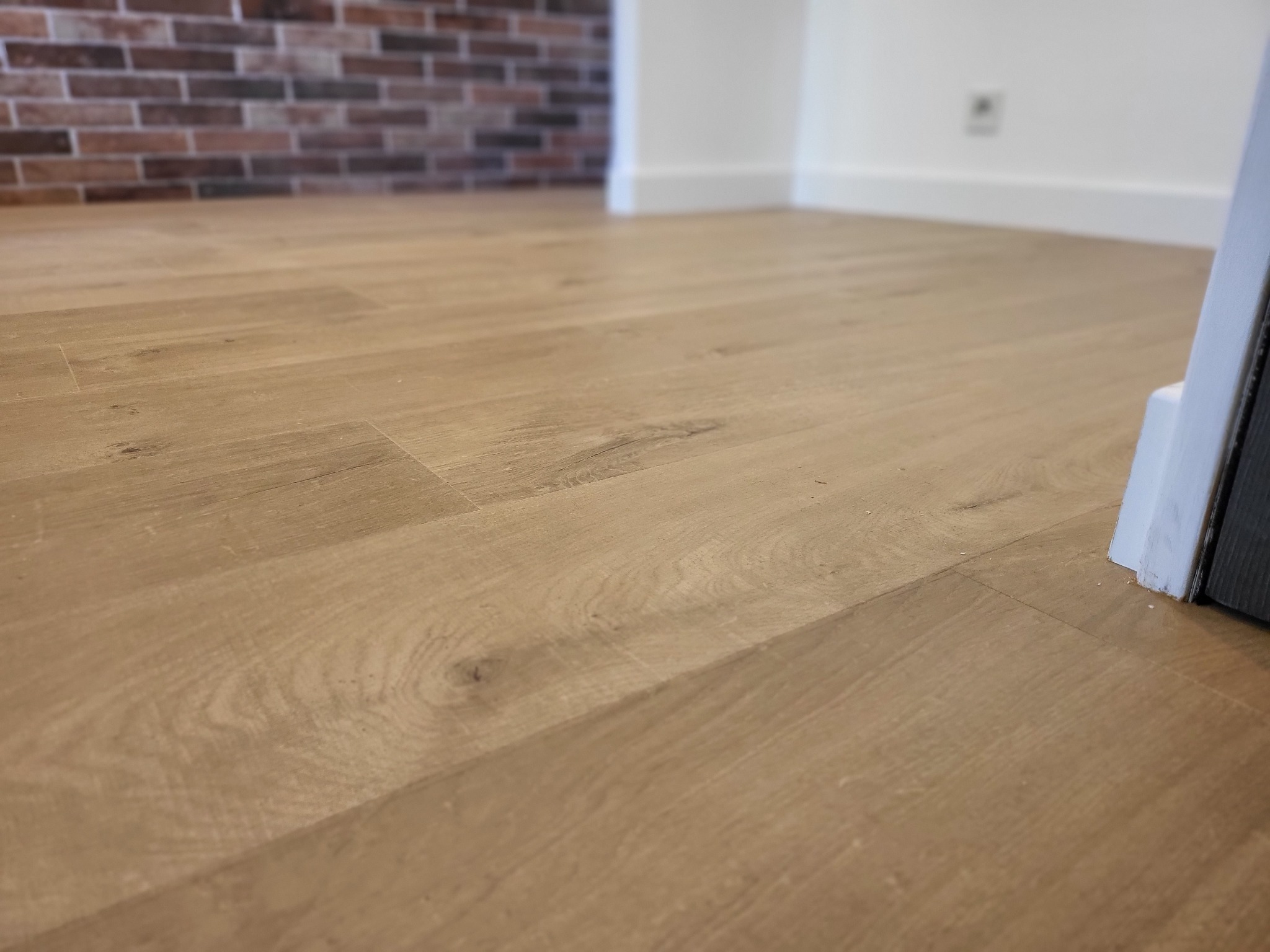 Ułożona jasna podłoga panelowa z widoczną strukturą drewna, w tle fragment ściany z cegły i biała listwa przypodłogowa