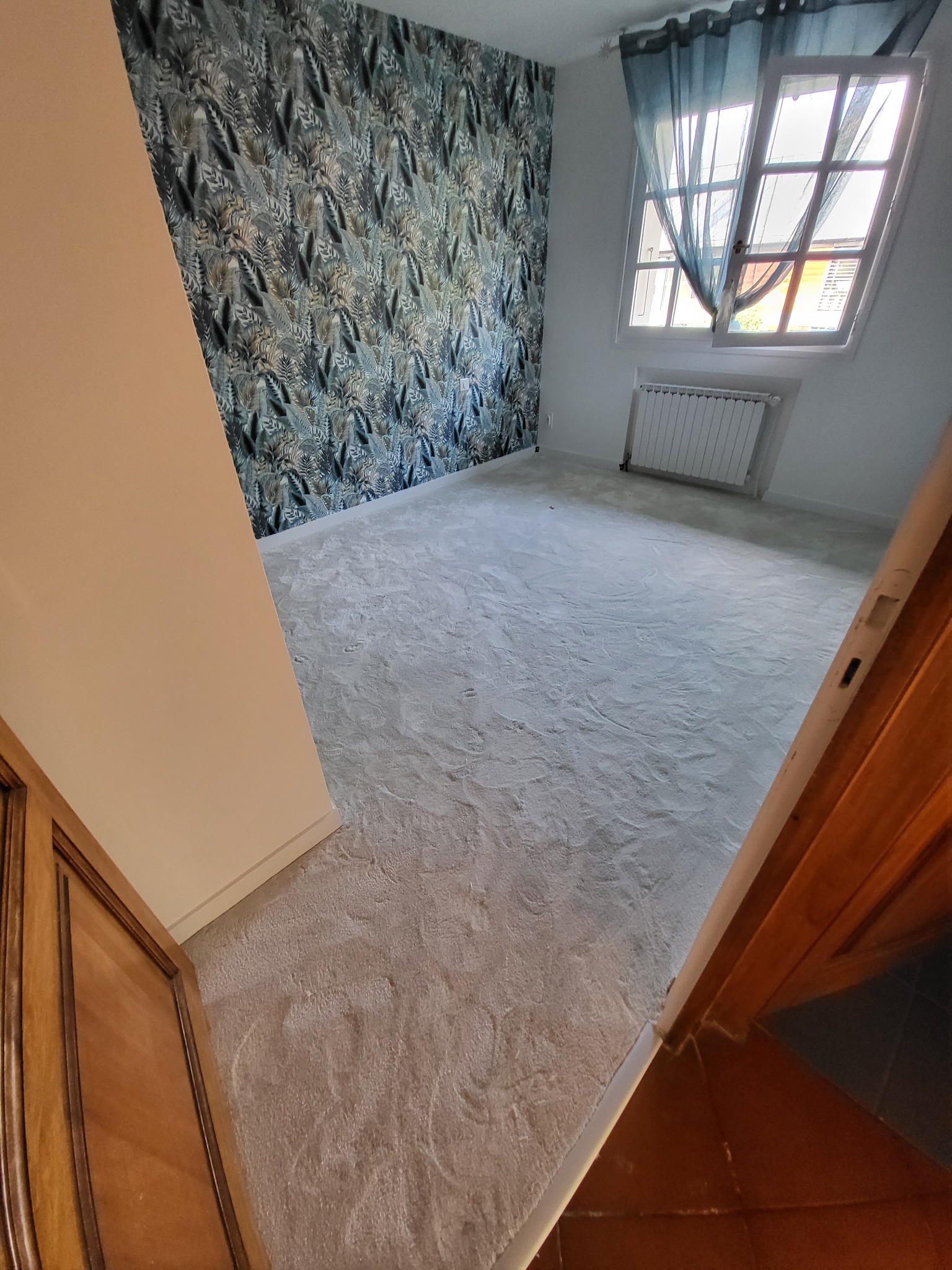 Pusty pokój po malowaniu ścian i położeniu nowej, jasnej wykładziny; widok zza uchylonych drewnianych drzwi, okno z firanką w tle.