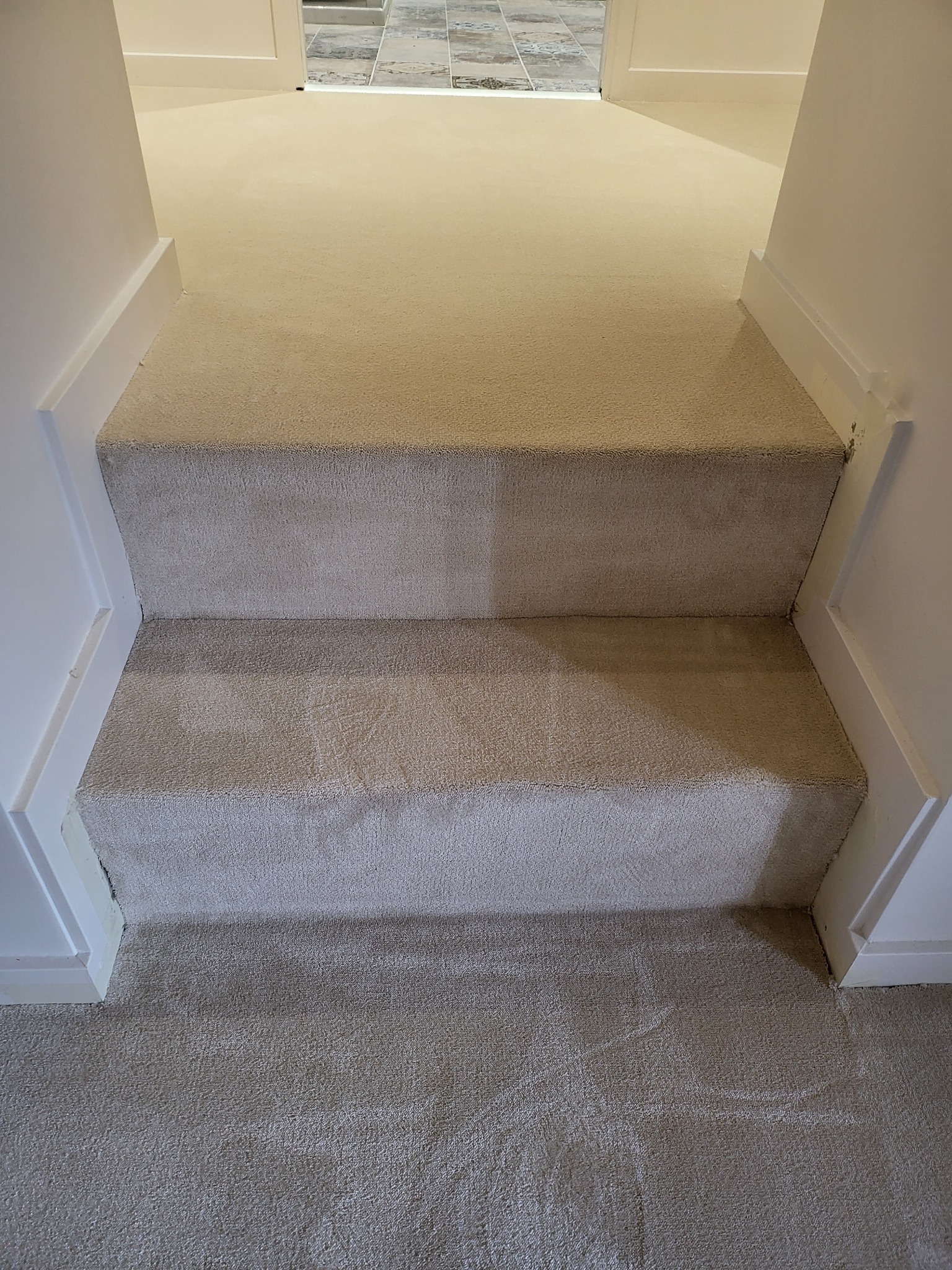 Dwa stopnie schodów pokryte beżową wykładziną dywanową, białe listwy przypodłogowe, widok z góry na korytarz z płytkami w tle.