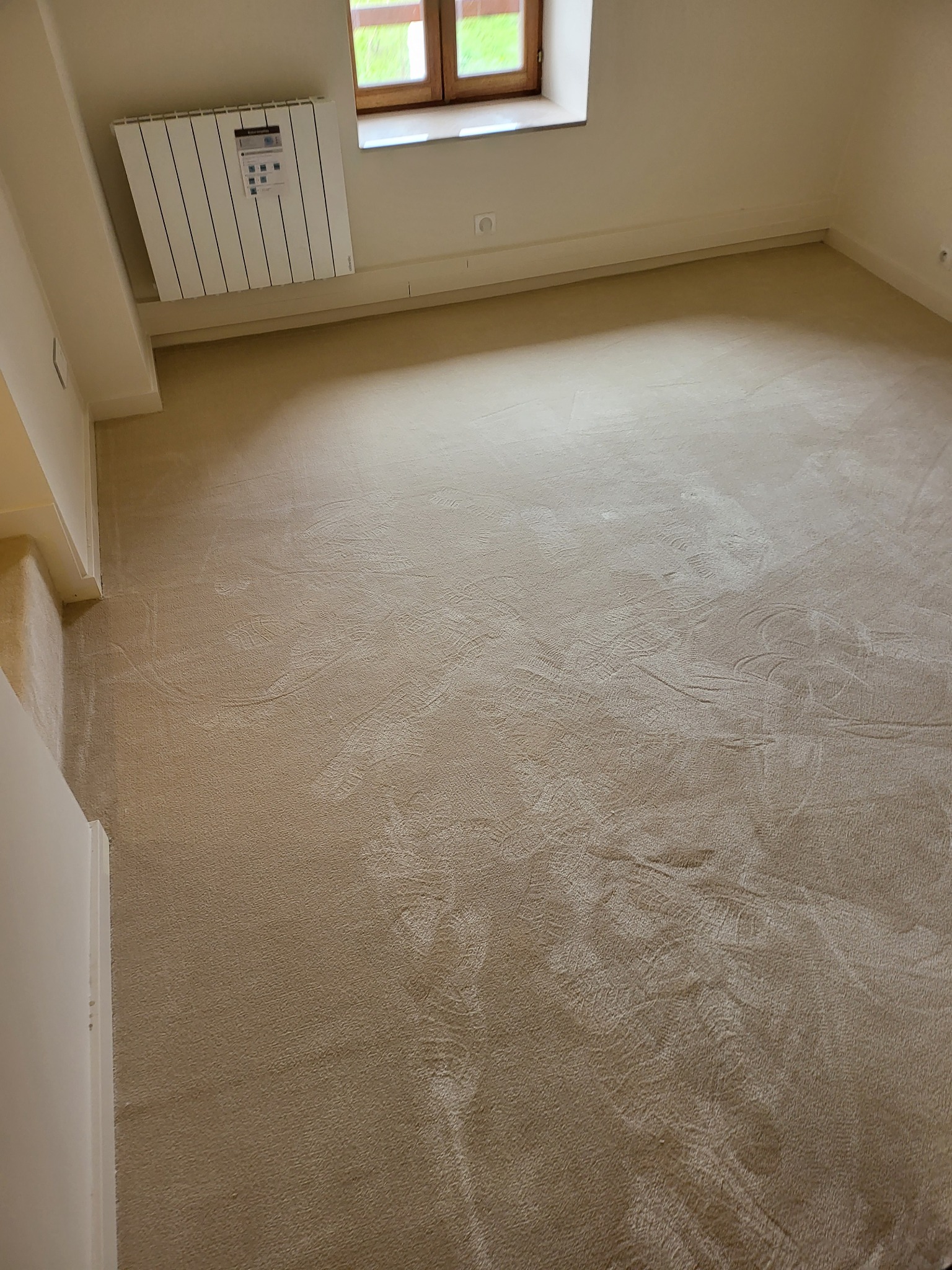Beżowa wykładzina dywanowa w pustym pokoju z grzejnikiem pod oknem, widoczne ślady czyszczenia