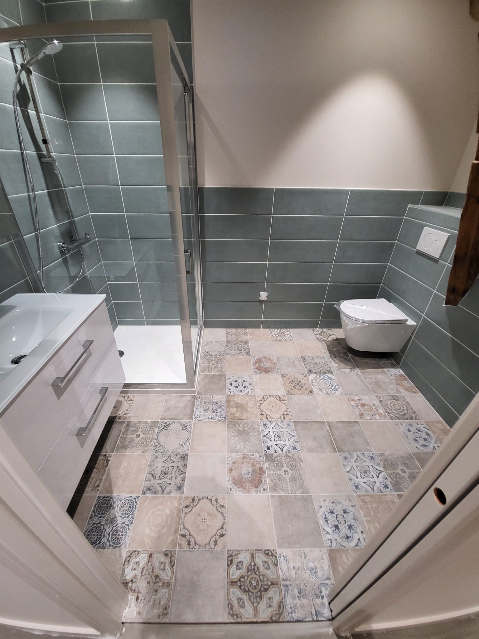 Nowoczesna łazienka z szarymi płytkami ściennymi, kabiną prysznicową, białą szafką pod umywalką i wzorzystą podłogą w stylu patchwork. Toaleta w folii ochronnej.