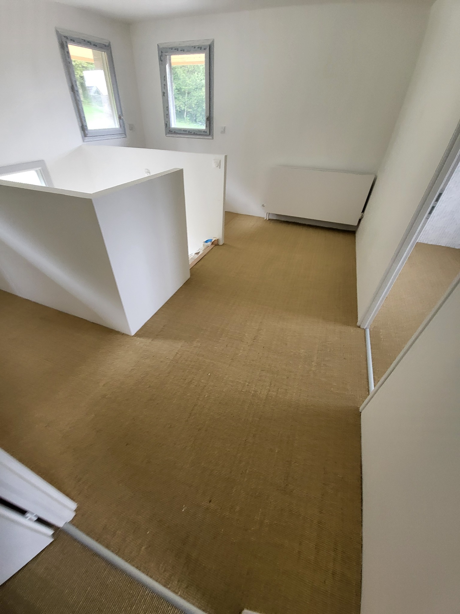 Wnętrze nowo wykończonego, jasnego pomieszczenia z beżową podłogą, białymi ścianami, grzejnikiem i oknami w szarych ramach.