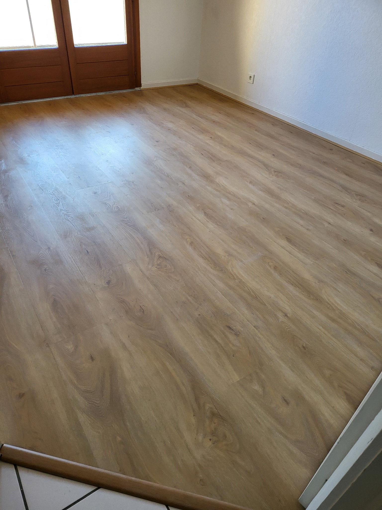 Jasny parkiet imitujący drewno ułożony w pustym pokoju z białymi ścianami i brązowymi drzwiami balkonowymi.