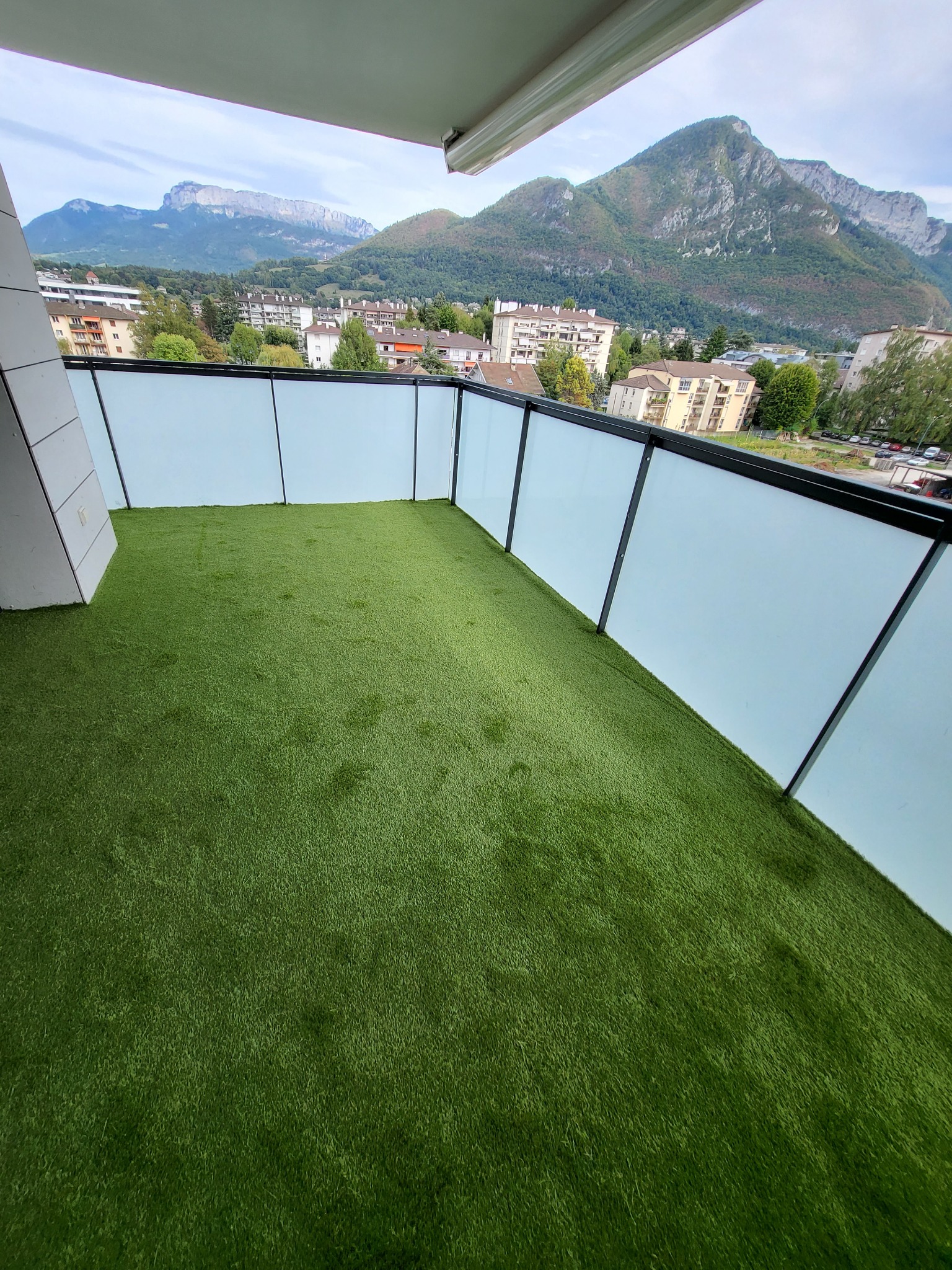 Wykończony balkon z zieloną sztuczną trawą, szklaną balustradą i widokiem na góry.