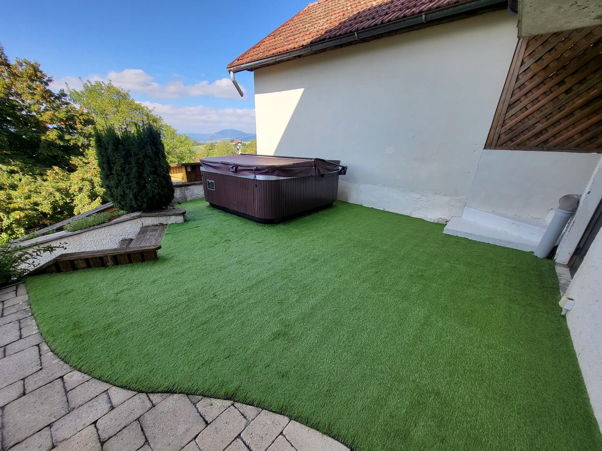 Brązowe jacuzzi z pokrywą na sztucznej trawie obok białego budynku z drewnianymi elementami, widok na wzgórza w tle.