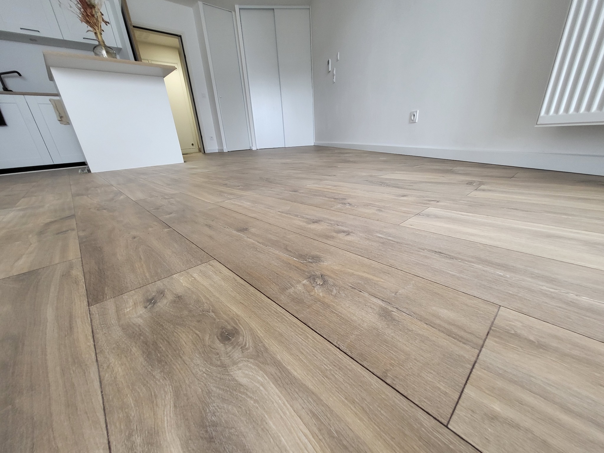 Jasne panele podłogowe imitujące drewno ułożone w nowym mieszkaniu, widok z niskiej perspektywy, z fragmentem białej zabudowy kuchennej i grzejnikiem w tle.