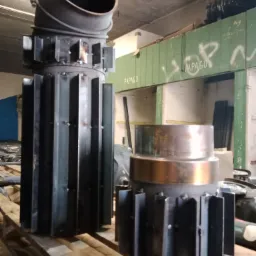 Dwa metalowe cylindryczne elementy konstrukcyjne z przyspawanymi pionowymi żebrami, jeden z ukośną rurą na górze, ustawione na drewnianych belkach w warsztacie.