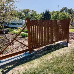 Przesuwna brama z pionowych, drewnianych szczebli w kolorze brązowym, zamontowana na betonowej podstawie z widocznym mechanizmem jezdnym, w tle zielony trawnik i drzewa.