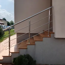 Zewnętrzne schody wyłożone terakotą z nowoczesną, minimalistyczną, srebrną poręczą z nierdzewnej stali, z budynkiem w tle.