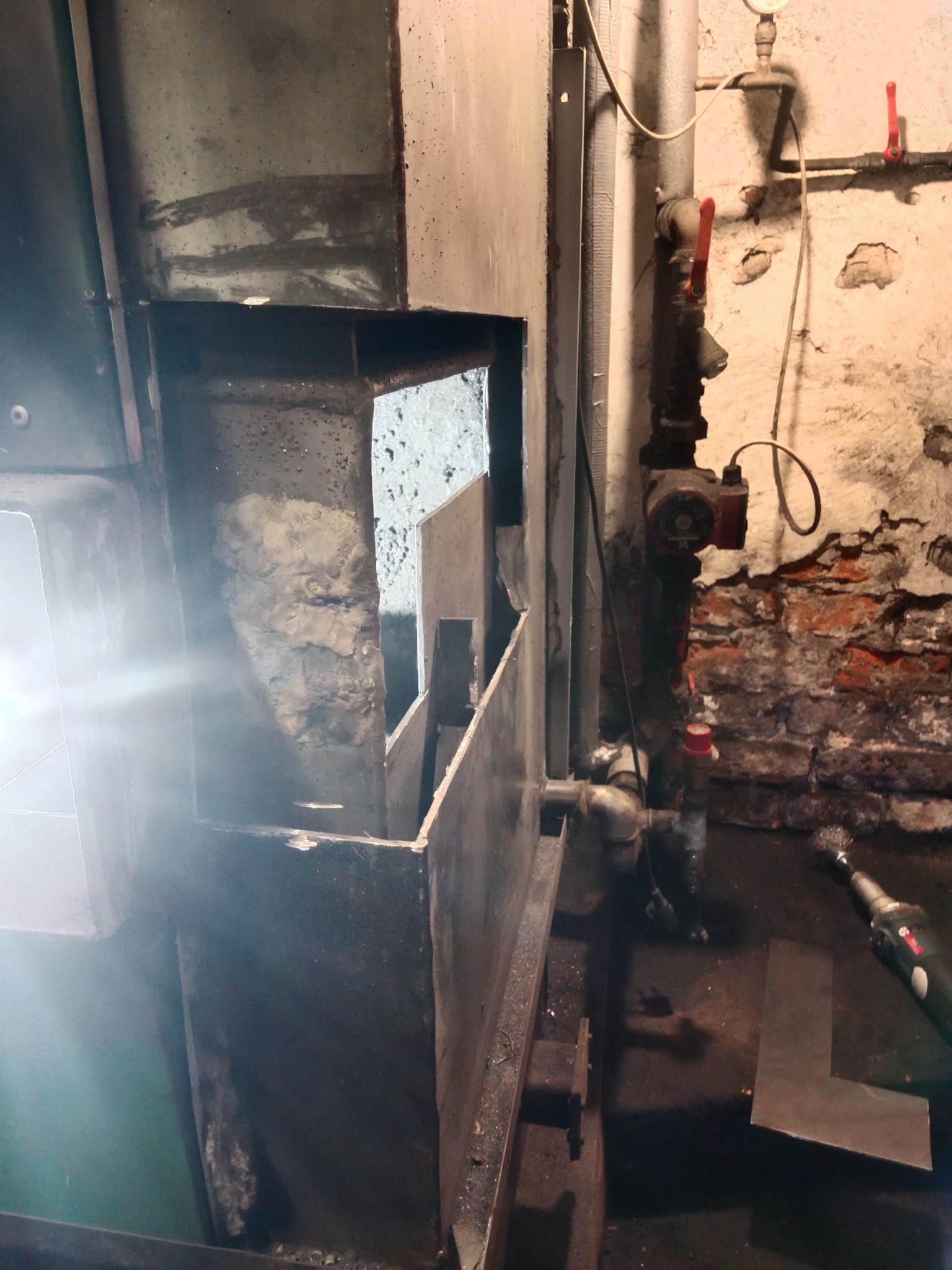 Fragment konstrukcji metalowej w trakcie obróbki, widoczne narzędzie szlifierskie i spawane elementy w pomieszczeniu z instalacją rurową na tle ściany z cegły