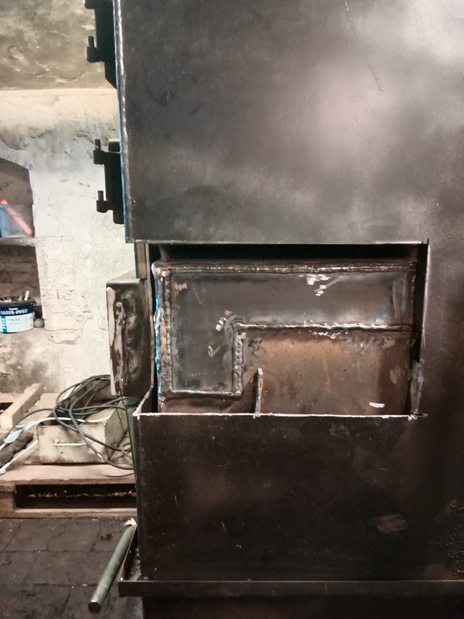 Fragment konstrukcji z grubych, spawanych elementów stalowych, widoczne spoiny, surowy stan wykończenia, w tle fragment pomieszczenia warsztatowego.