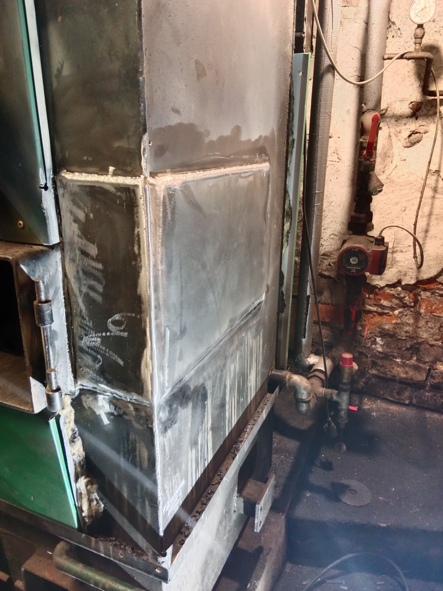 Metalowa konstrukcja pieca centralnego ogrzewania w trakcie naprawy, widoczne ślady spawania i obróbki metalu, instalacja hydrauliczna w tle.