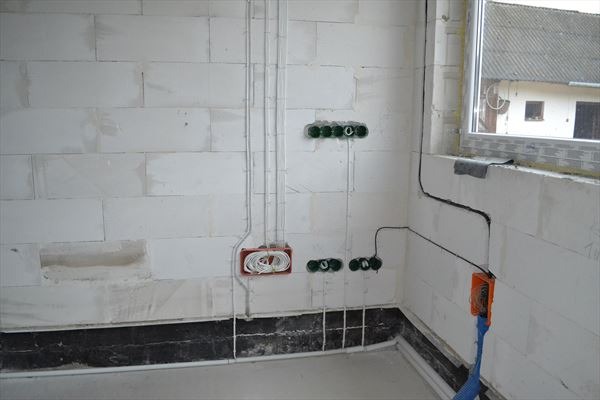 Surowe wnętrze z białego bloczka, widoczne puszki instalacyjne z przewodami elektrycznymi, podłączenia pod okno, instalacja w trakcie wykonywania.