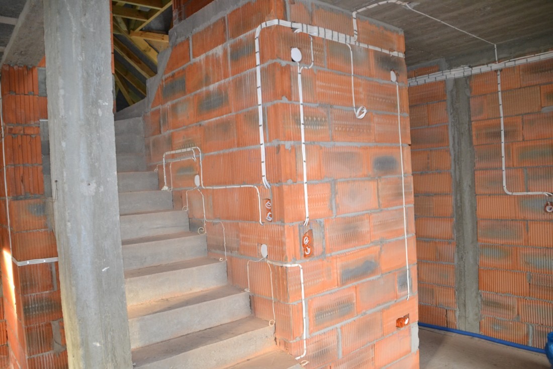 Wewnętrzna instalacja przewodów elektrycznych w białych listwach na ścianie z czerwonej cegły dziurawki w nowo budowanym domu, widoczne schody betonowe.