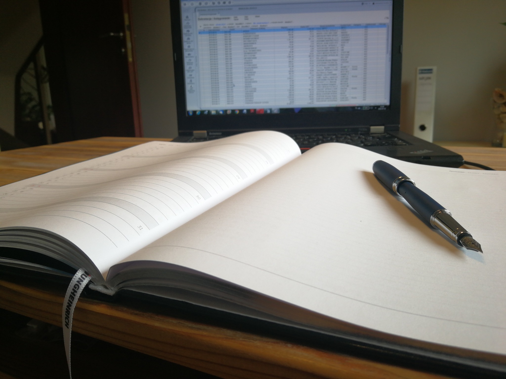 Otwarty kalendarz leżący na drewnianym biurku, obok pióro wieczne i laptop z widocznym arkuszem kalkulacyjnym. W tle widoczny fragment pomieszczenia biurowego.