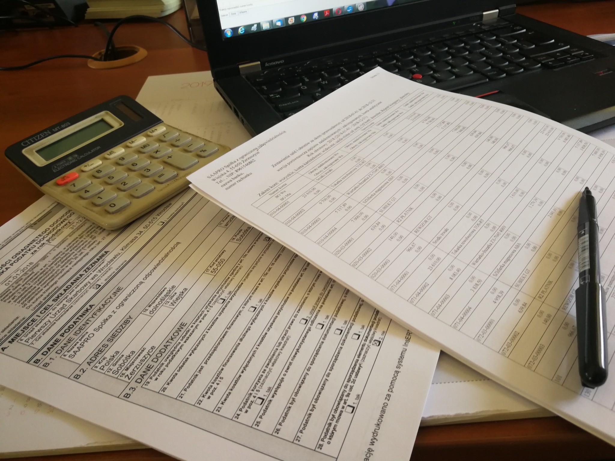 Formularze podatkowe, kalkulator i długopis na biurku z laptopem w tle, ukazujące pracę biurową.