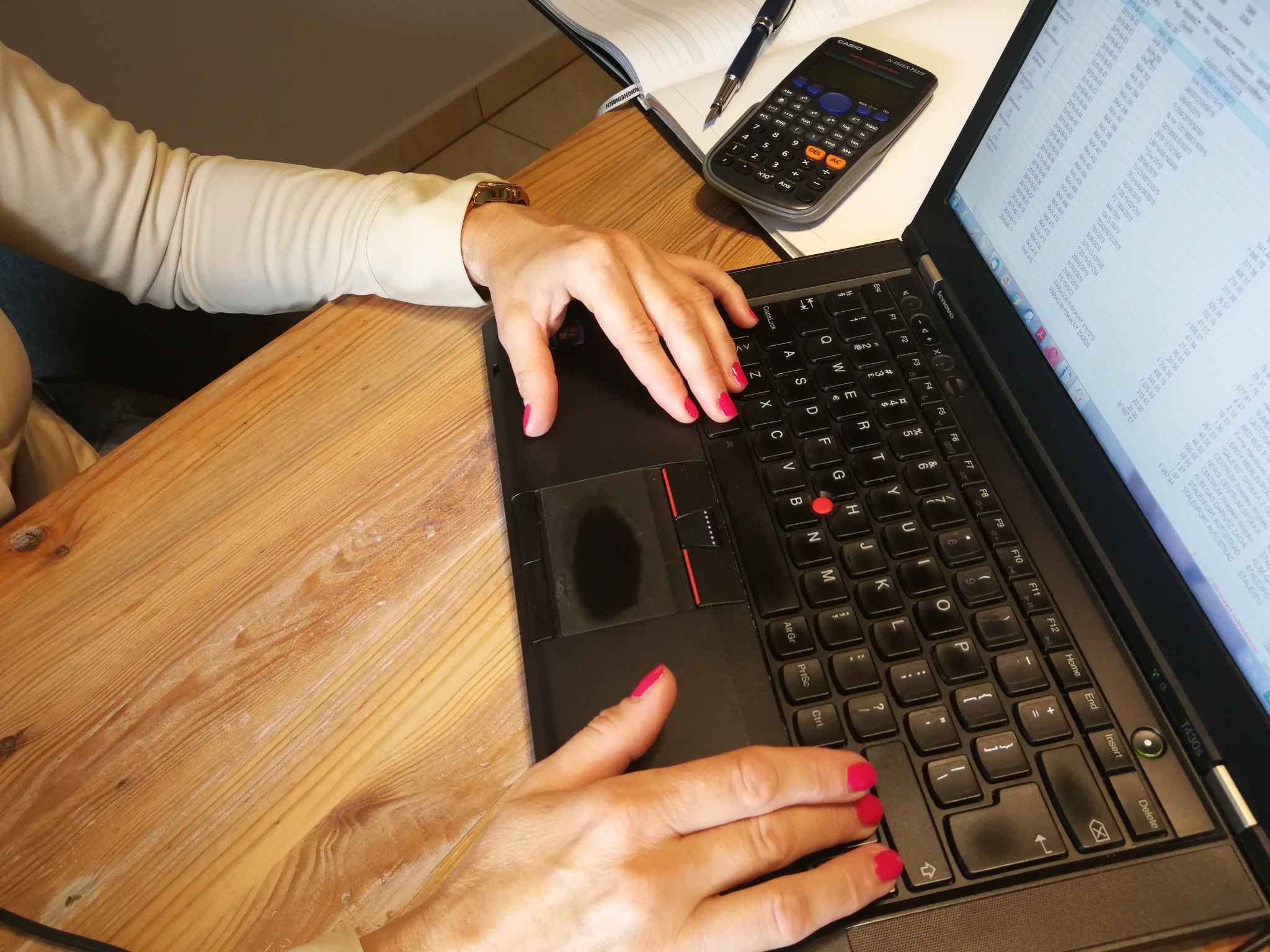 Kobieta z różowym manicure pracuje na laptopie, obok kalkulator, notatnik i długopis, widoczne dane na ekranie.