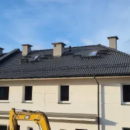 Nowoczesny dom z ciemną dach&oacute;wką karpi&oacute;wką, lukarnami i czterema kominami o kwadratowym przekroju, z częściowo widoczną koparką CAT w dolnej części kadru.