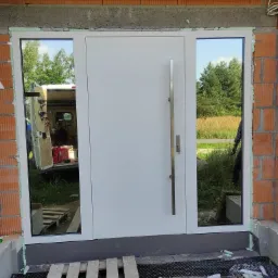 Białe drzwi wejściowe z bocznymi przeszkleniami, zamontowane w surowym stanie budynku, widoczne narzędzia i samochód dostawczy w odbiciu szyb.