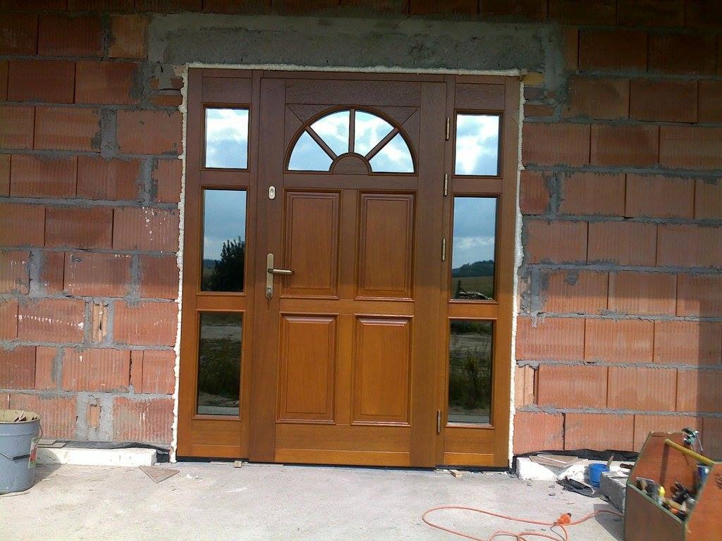 Brązowe drzwi wejściowe z szybami bocznymi i półokrągłym przeszkleniem u góry, zamontowane w surowym murze z czerwonej cegły, widoczne narzędzia i wiadro na podłodze.