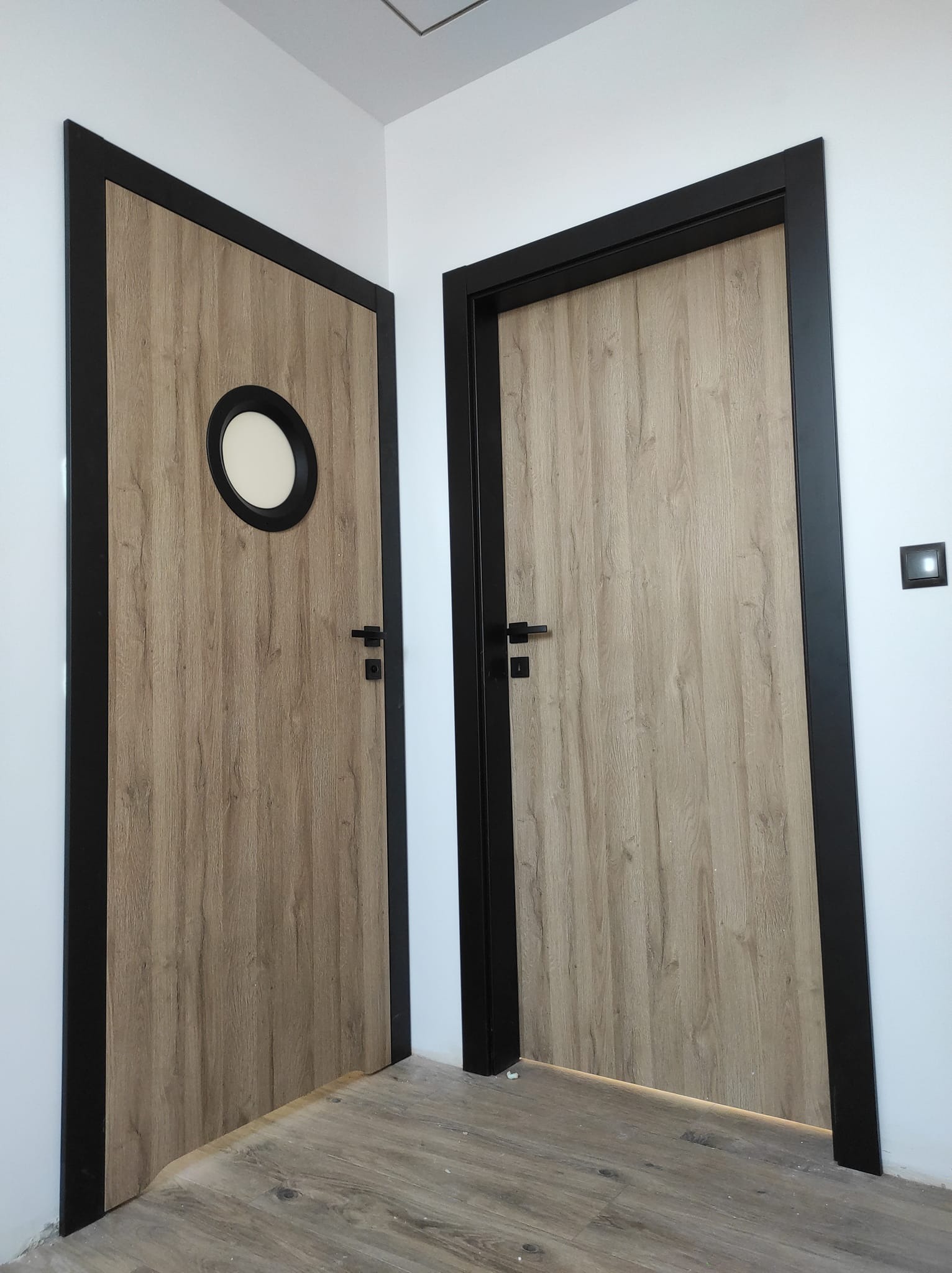 Dwa drewnopodobne skrzydła drzwiowe z czarnymi ościeżnicami, jedno z okrągłym okienkiem, zamontowane w białym pomieszczeniu.