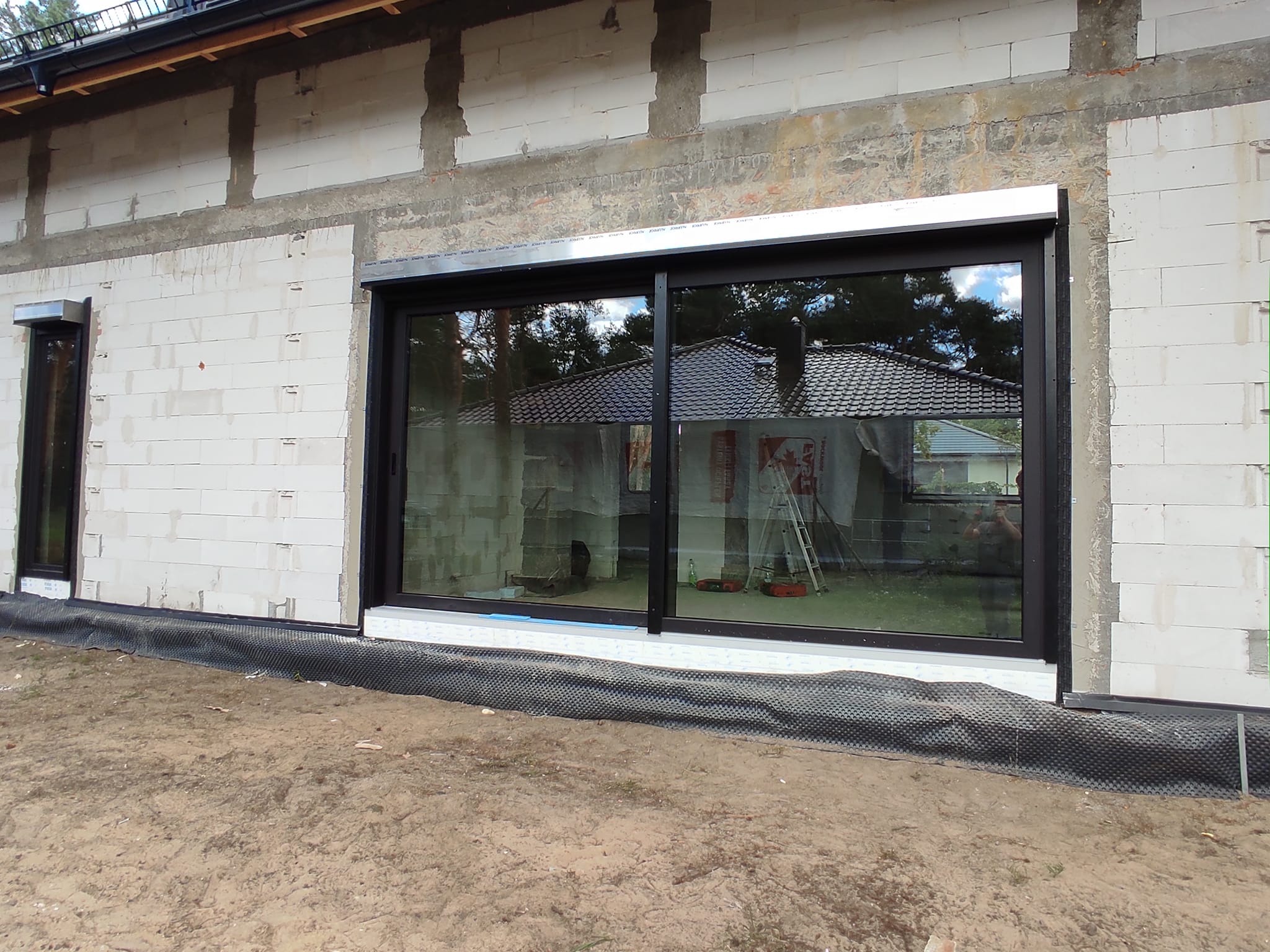 Montaż okna PCV w surowym budynku z białego pustaka, widoczne rolety zewnętrzne w kolorze ramy okiennej i czarna folia fundamentowa.