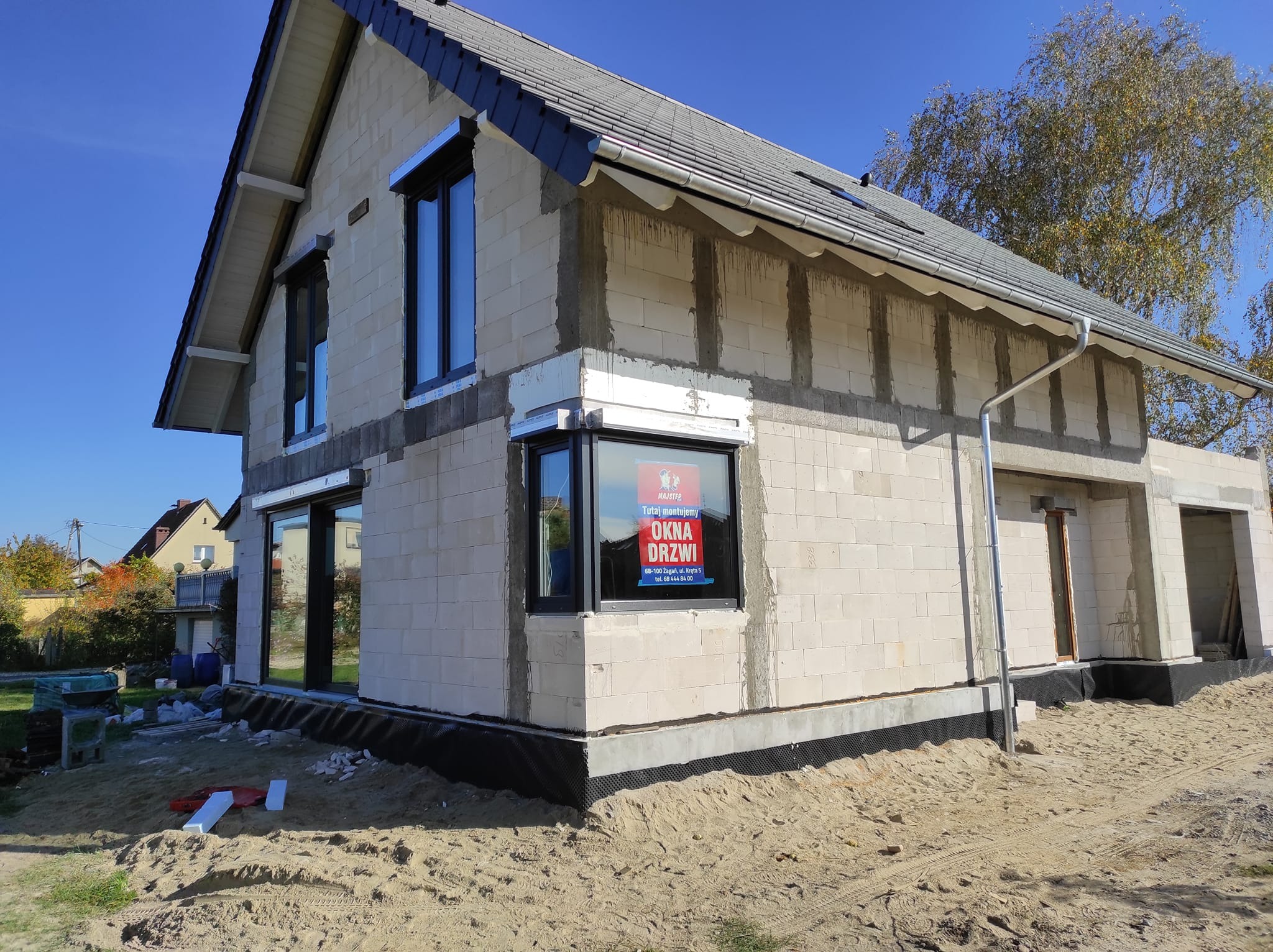 Budowa domu jednorodzinnego w trakcie montażu stolarki okiennej, widoczne okna z czarnymi ramami i szyld reklamowy firmy montującej okna i drzwi, tło stanowi błękitne niebo i drzewo.