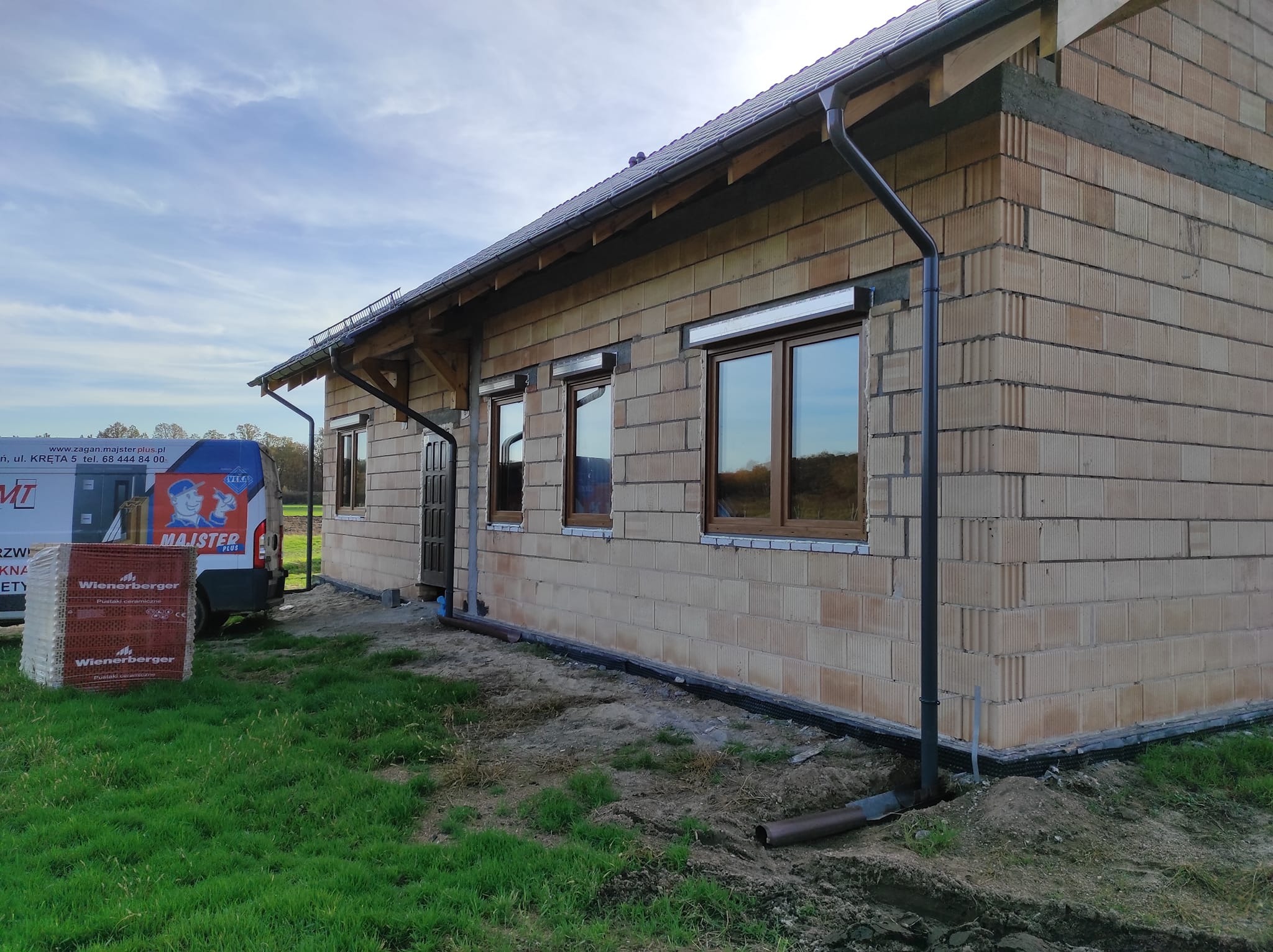 Surowy budynek z beżowych pustaków z zamontowanymi brązowymi oknami PCV i zwiniętymi roletami zewnętrznymi, widoczny samochód dostawczy firmy i paleta pustaków Wienerberger.