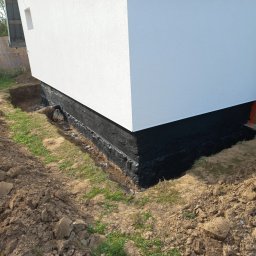 Patryk - Biała ściana budynku z czarną izolacją fundamentu, widoczny wykop ziemny. Detal zabezpieczenia przed wilgocią, świeżo po wykonaniu, z ziemią wokół.