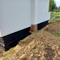 Patryk - Biała elewacja budynku z czarną hydroizolacją fundamentów, obok wykopana ziemia i narzędzia ogrodnicze. Widok z zewnątrz, w tle zieleń drzew.