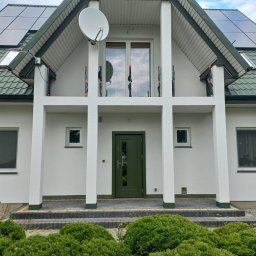 Patryk - Biała fasada domu z zielonym dachem, panelami fotowoltaicznymi i ciemnozielonymi drzwiami wejściowymi. Nad wejściem balkon z kolumnami i ozdobnymi, czarnymi balustradami.