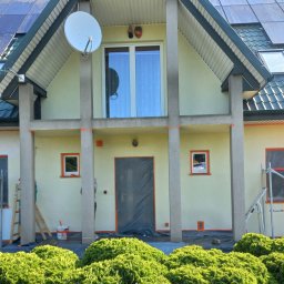 Patryk - Jasnożółta elewacja domu z zielonym dachem i panelami słonecznymi, w trakcie odnawiania. Okna i drzwi zabezpieczone folią i pomarańczową taśmą, widoczne rusztowanie i numer '1c'.
