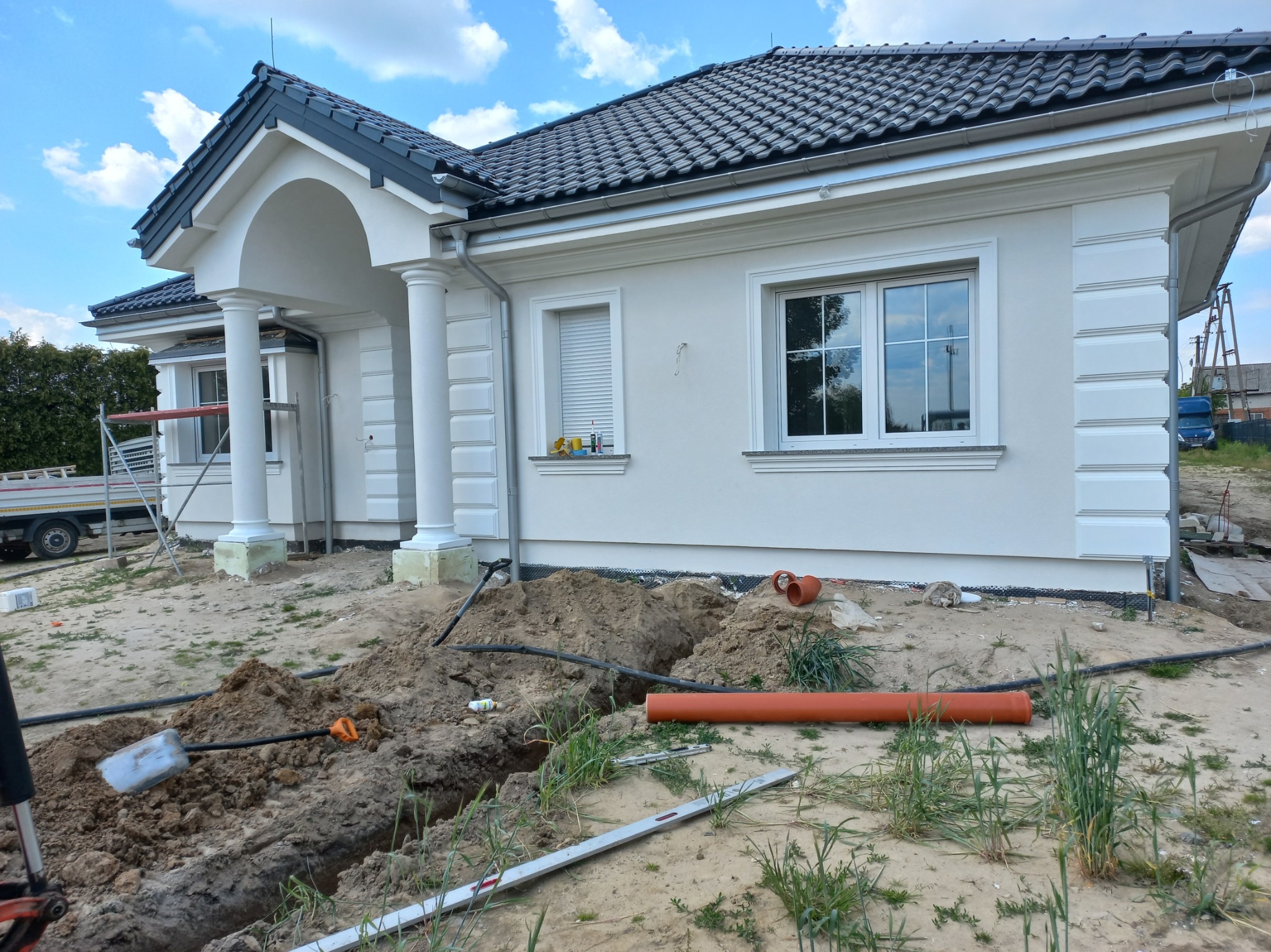 Elewacja domu jednorodzinnego w trakcie prac ziemnych. Widoczne rury, wykop i narzędzia. Biała fasada z kolumnami i szarą dachówką. Błękitne niebo w tle.