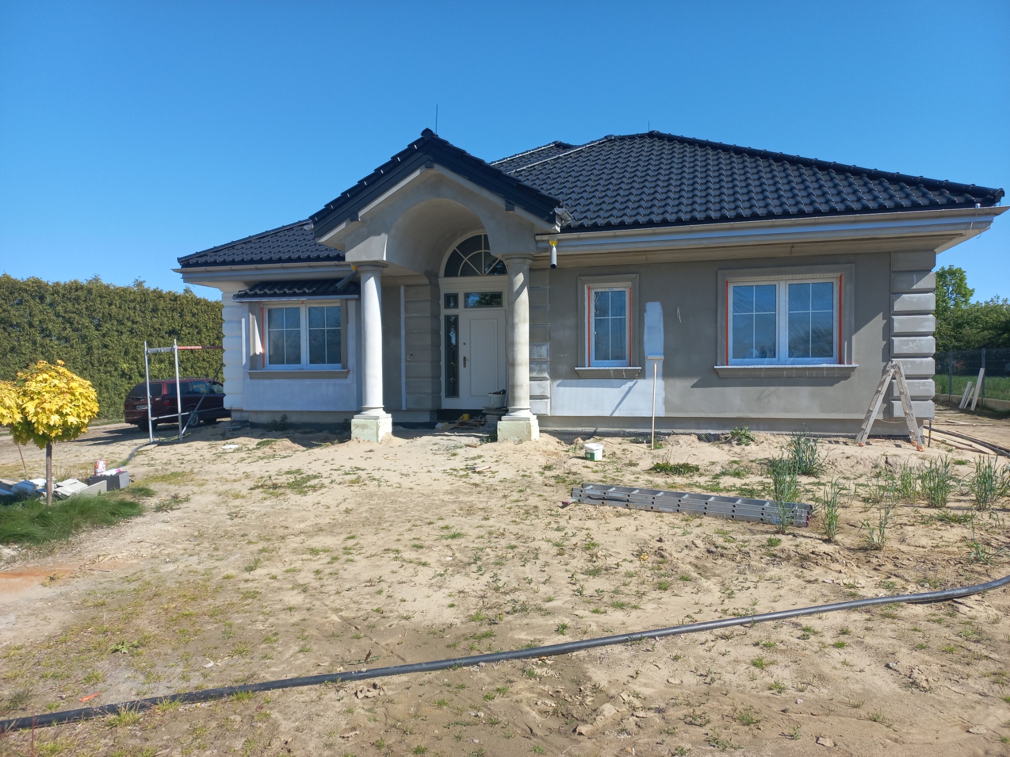 Dom jednorodzinny z ciemnym dachem i jasną elewacją w trakcie prac wykończeniowych. Widoczne rusztowania i narzędzia. Błękitne niebo w tle.