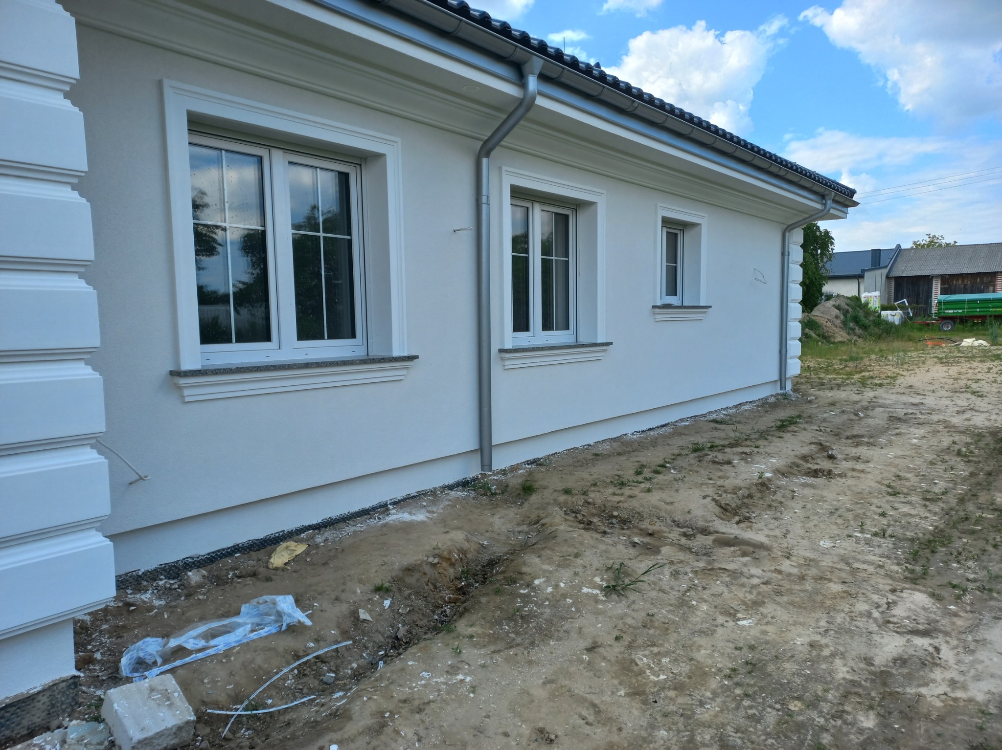 Biała elewacja domu z trzema oknami o różnych rozmiarach, ozdobnymi ramami i gzymsami. Widoczny fragment dachu z rynnami i teren wokół budynku w trakcie prac ziemnych.