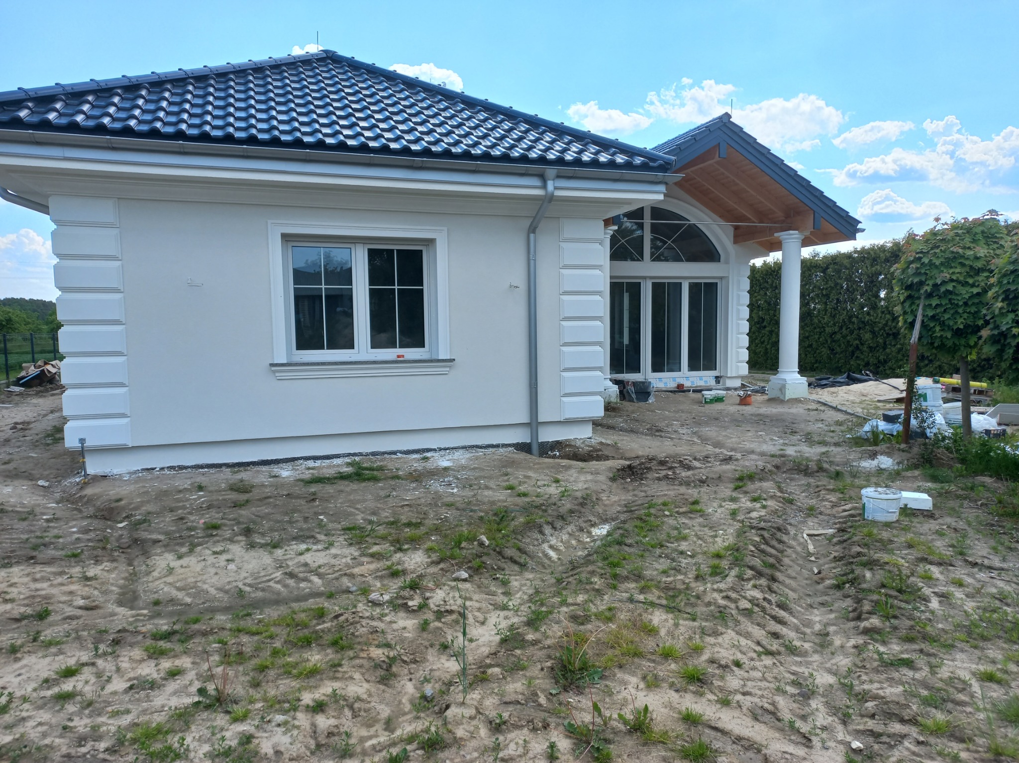 Biała elewacja domu jednorodzinnego z dekoracyjnymi elementami sztukaterii, widok z zewnątrz, teren wokół domu w trakcie prac wykończeniowych.