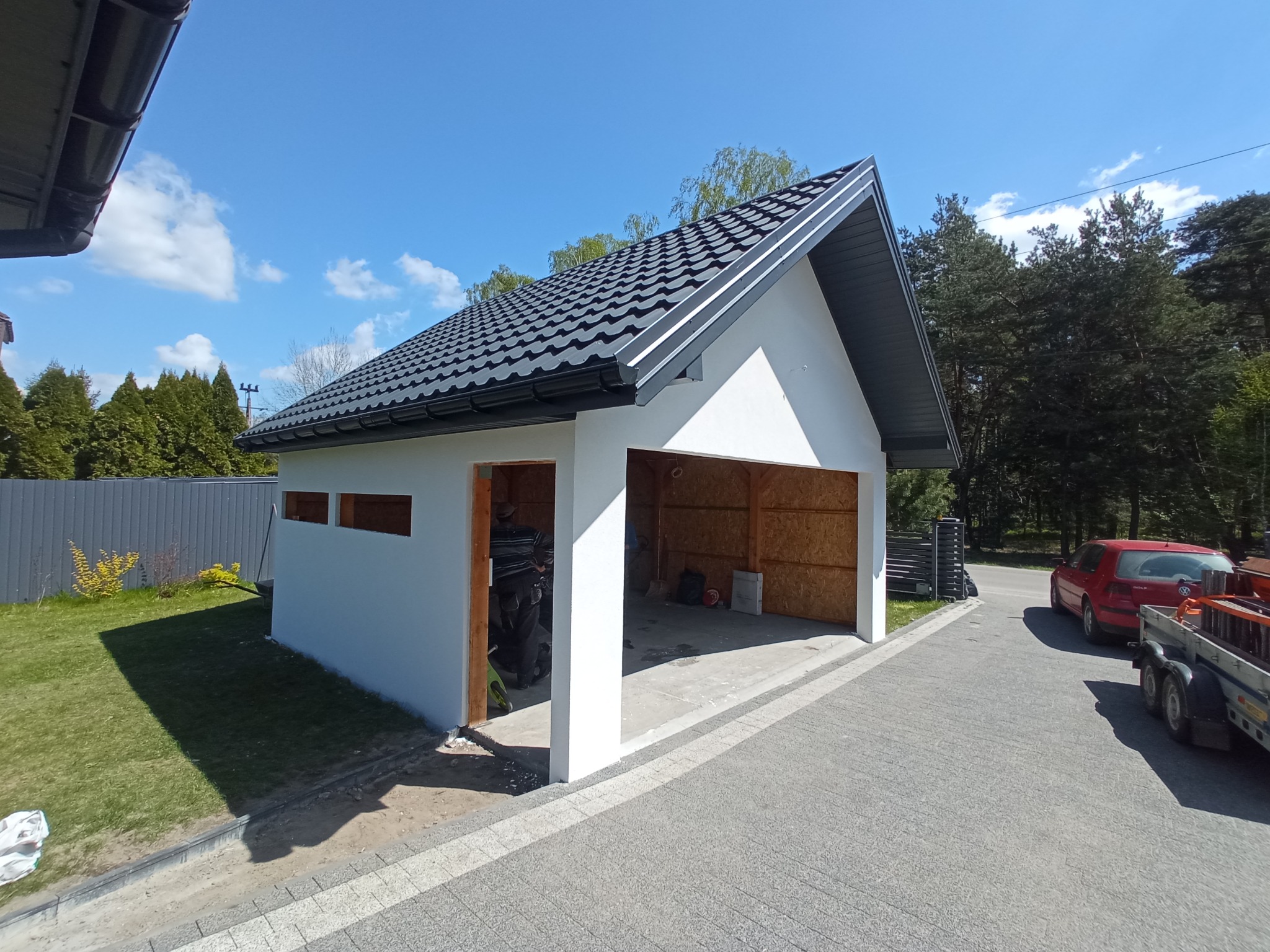 Nowoczesny, biały garaż z czarnym, dwuspadowym dachem z blachodachówki, widoczny z zewnątrz, z podjazdem wyłożonym kostką brukową i zielenią w tle.
