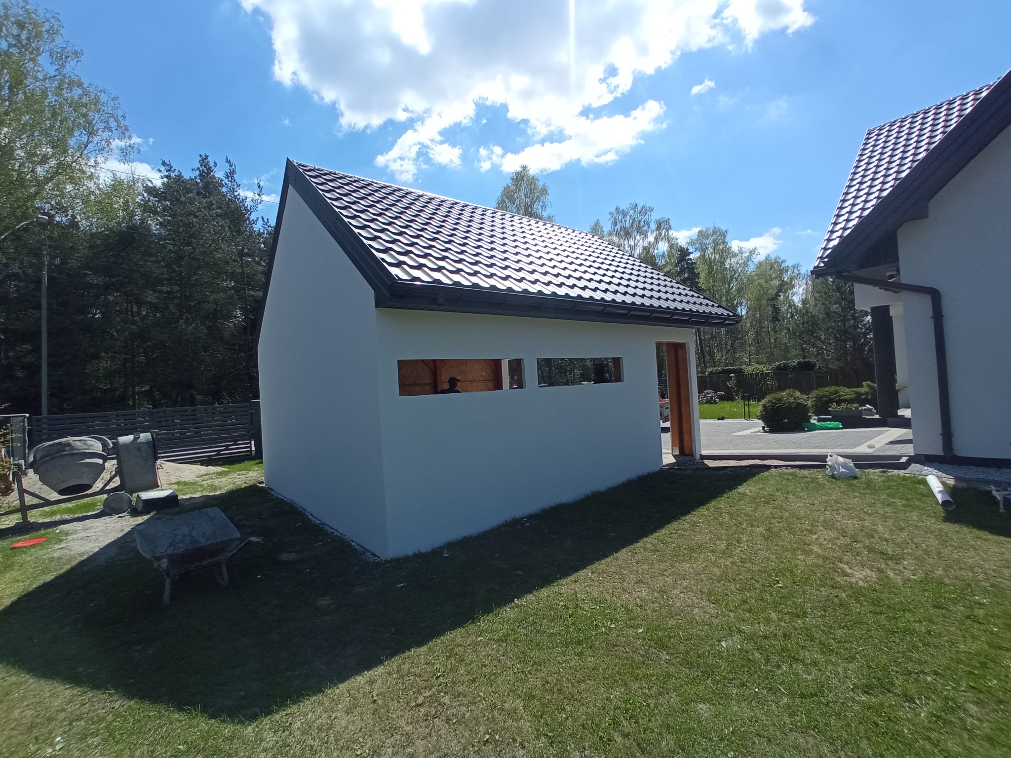 Nowo wybudowany, biały budynek gospodarczy z ciemną, metalową blachodachówką w słoneczny dzień. Widoczne trzy okna bez szyb, otwarte drzwi i maszyna do betonu w tle.