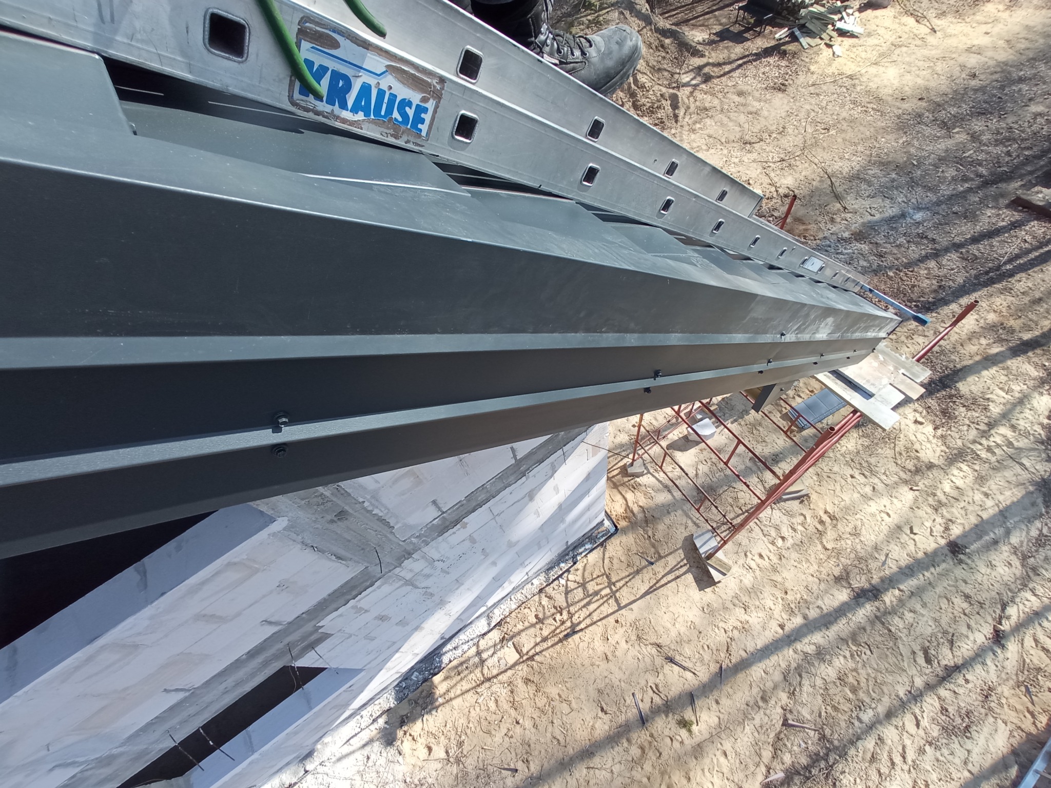 Widok z góry na fragment dachu pokrytego szarą blachą, z drabiną Krause opartą o jego krawędź, w tle surowa ściana budynku z betonu komórkowego i piaszczyste podłoże.