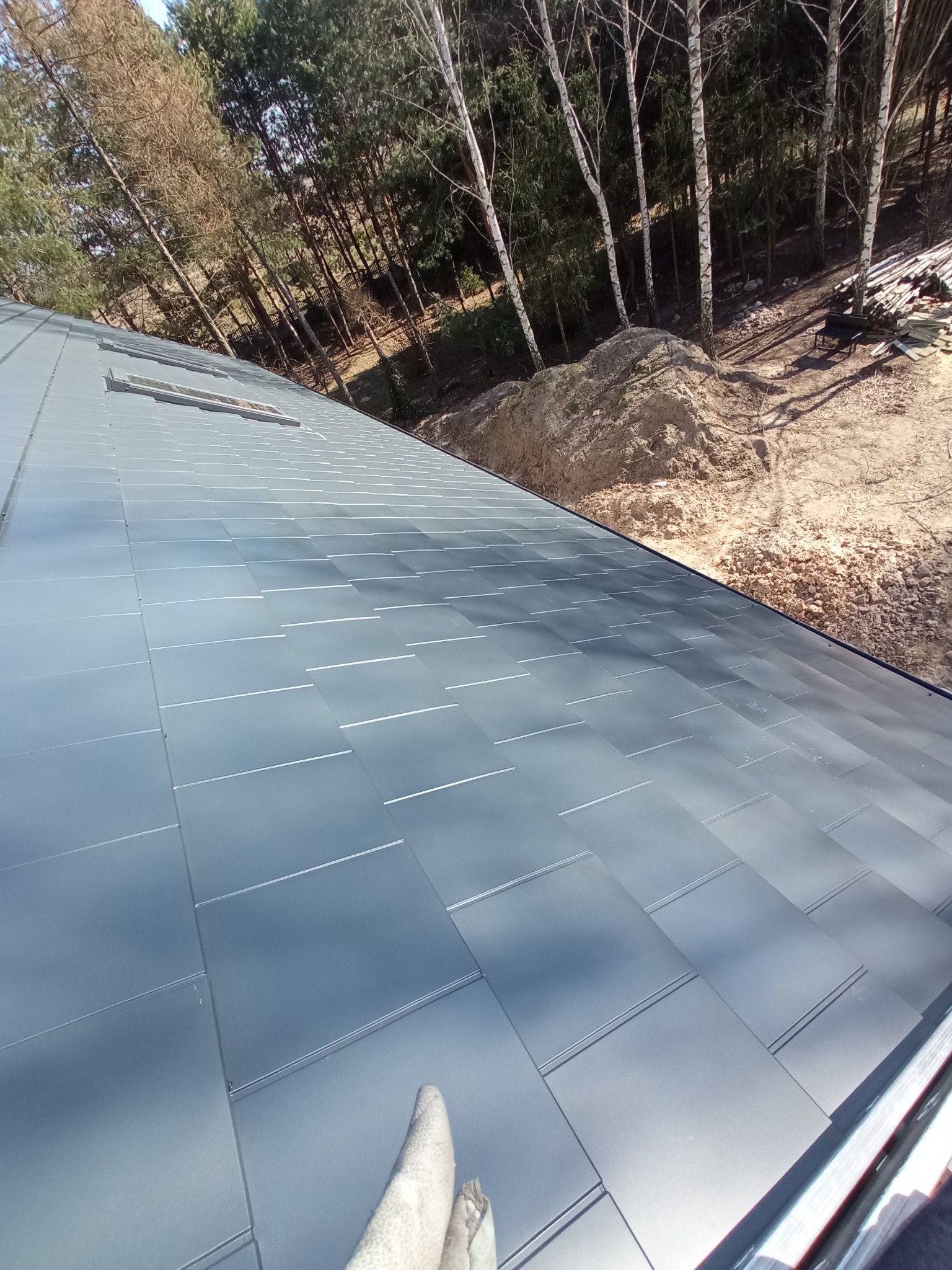 Nowo położony, grafitowy dach z blachy na tle lasu i ziemi, widoczny fragment rękawicy roboczej w dolnym rogu kadru.