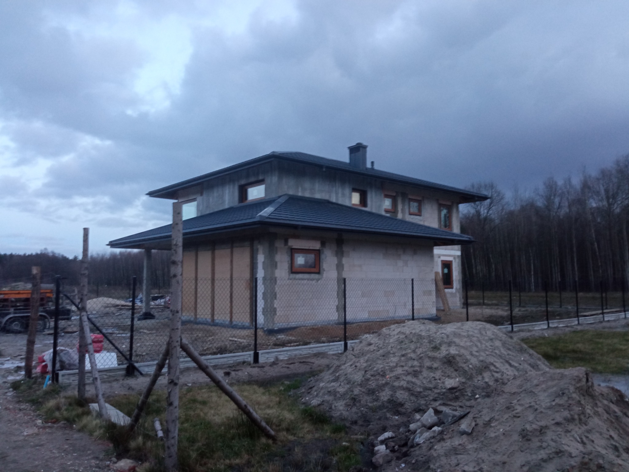 Dom w trakcie budowy, z widoczną konstrukcją dachu pokrytą ciemną dachówką, ściany z bloczków, teren budowy ogrodzony siatką, pochmurne niebo.