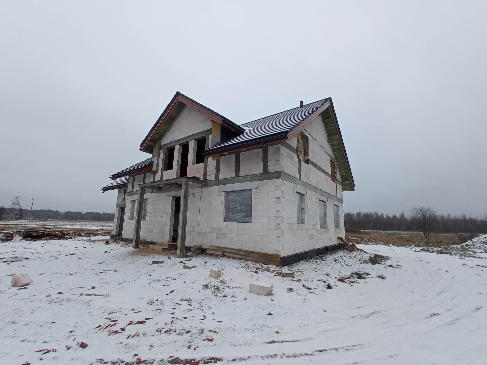 Dom w stanie surowym otwartym z częściowo ukończonym dachem w Ulan-Majorat, widoczne fragmenty więźby dachowej i czarna dachówka na tle zimowego krajobrazu.