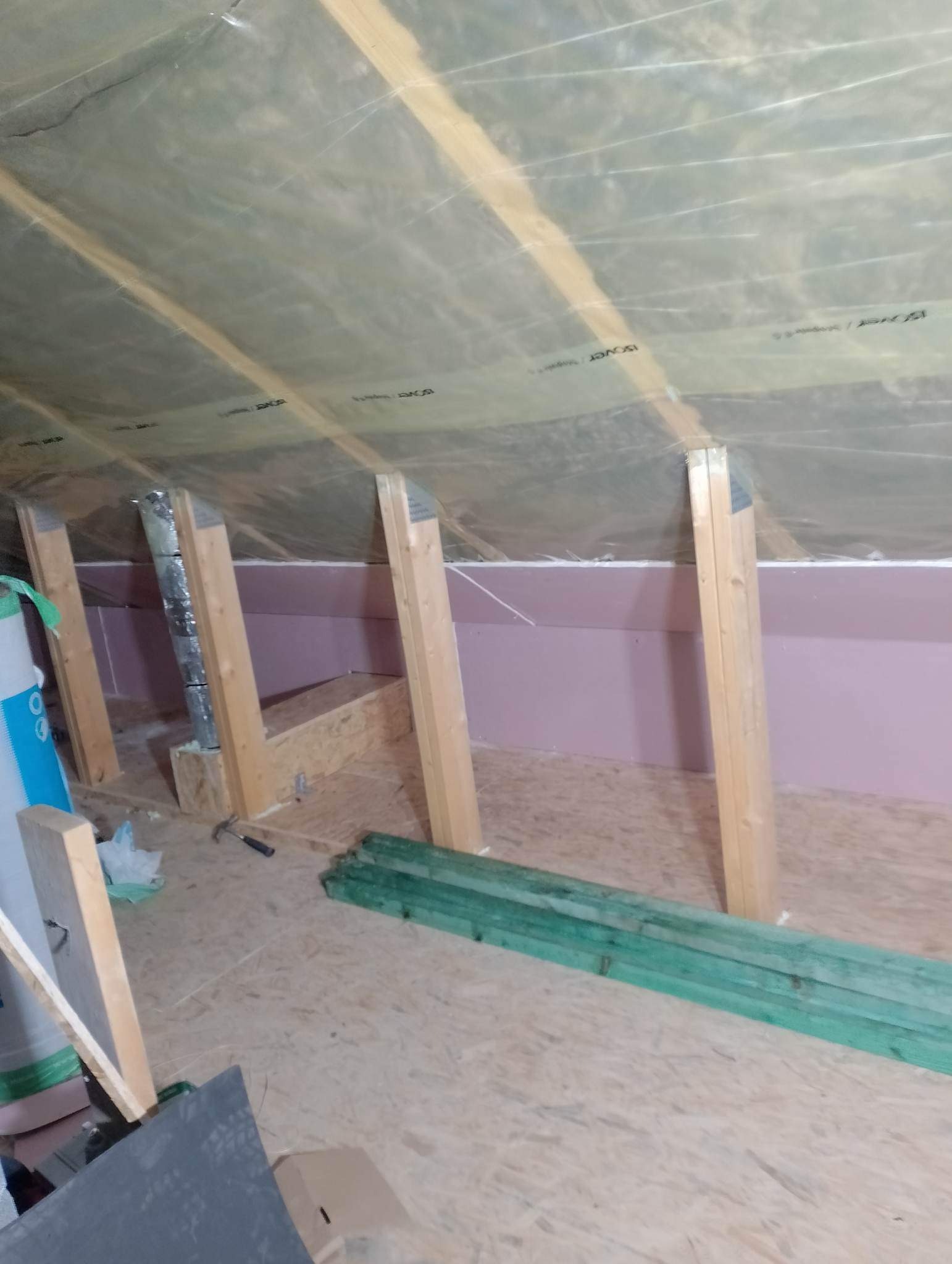 Ocieplenie poddasza wełną mineralną, widoczne krokwie, folia paroizolacyjna i płyty styropianowe na ścianach kolankowych, przygotowane zielone deski na podłodze.