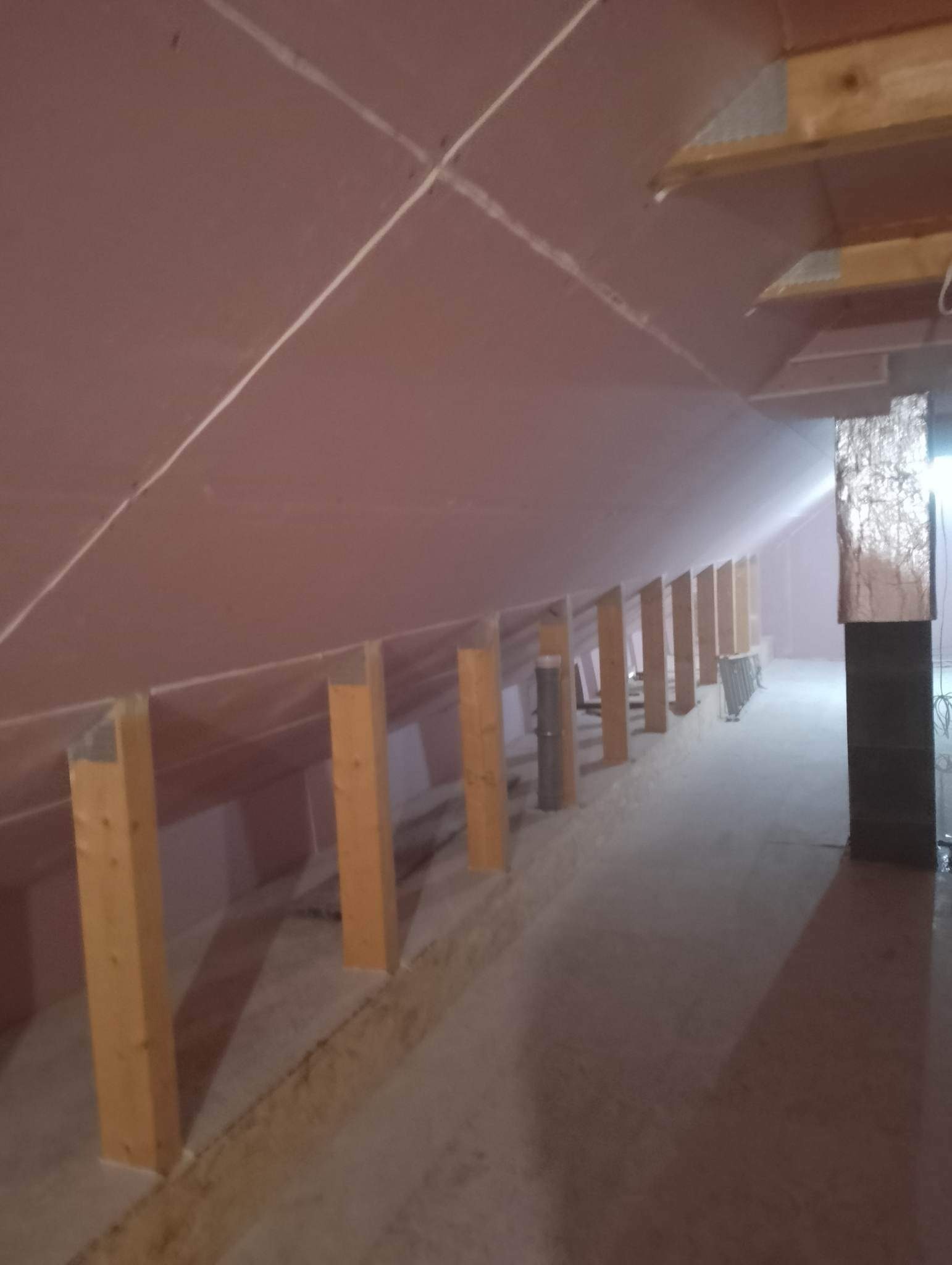Poddasze w trakcie ocieplania: widoczne drewniane słupki konstrukcyjne, wełna mineralna na skosach dachu i fragment komina w izolacji aluminiowej.