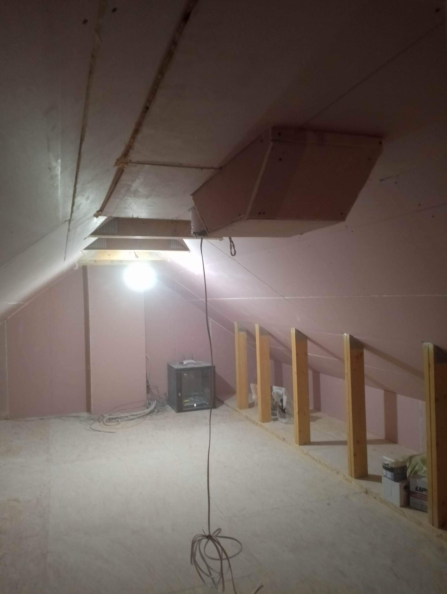Poddasze w trakcie adaptacji: ściany wyłożone płytami kartonowo-gipsowymi w kolorze różowym, widoczne drewniane słupki konstrukcyjne, wystająca instalacja wentylacyjna i przewody elektryczne...