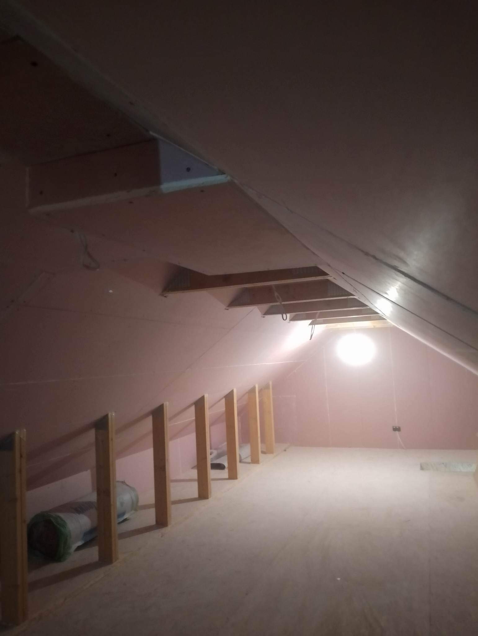 Poddasze w trakcie adaptacji: widoczne drewniane słupki, ocieplone ściany płytami gipsowo-kartonowymi i instalacja elektryczna, z rolką materiału izolacyjnego leżącą na podłodze.