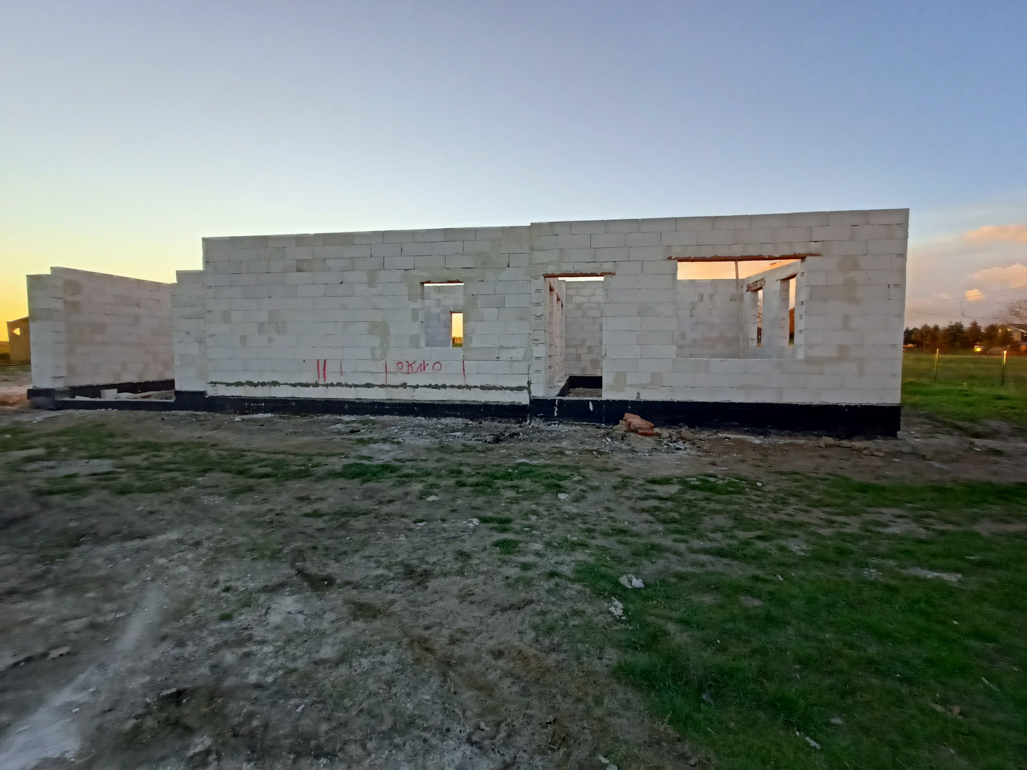 Surowy stan domu jednorodzinnego w trakcie budowy z białych bloczków, widoczne oznaczenia 'OKNO' czerwoną farbą, czarna izolacja fundamentów i zarys zachodzącego słońca w oknach.