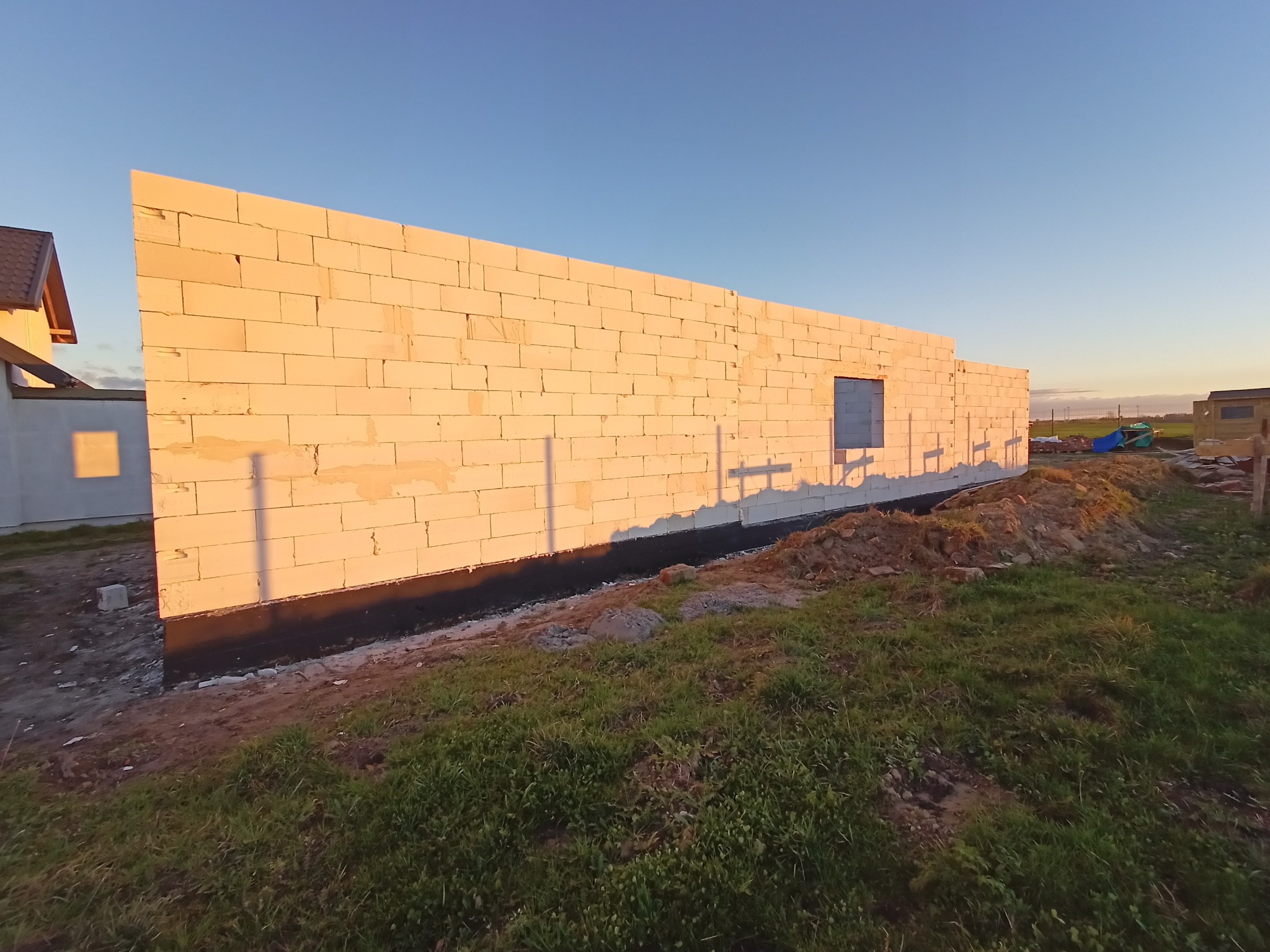 Ściana domu w trakcie budowy, wzniesiona z białych bloczków, zabezpieczona czarną izolacją przeciwwilgociową u podstawy, w tle fragment dachu sąsiedniego budynku i błękitne niebo.