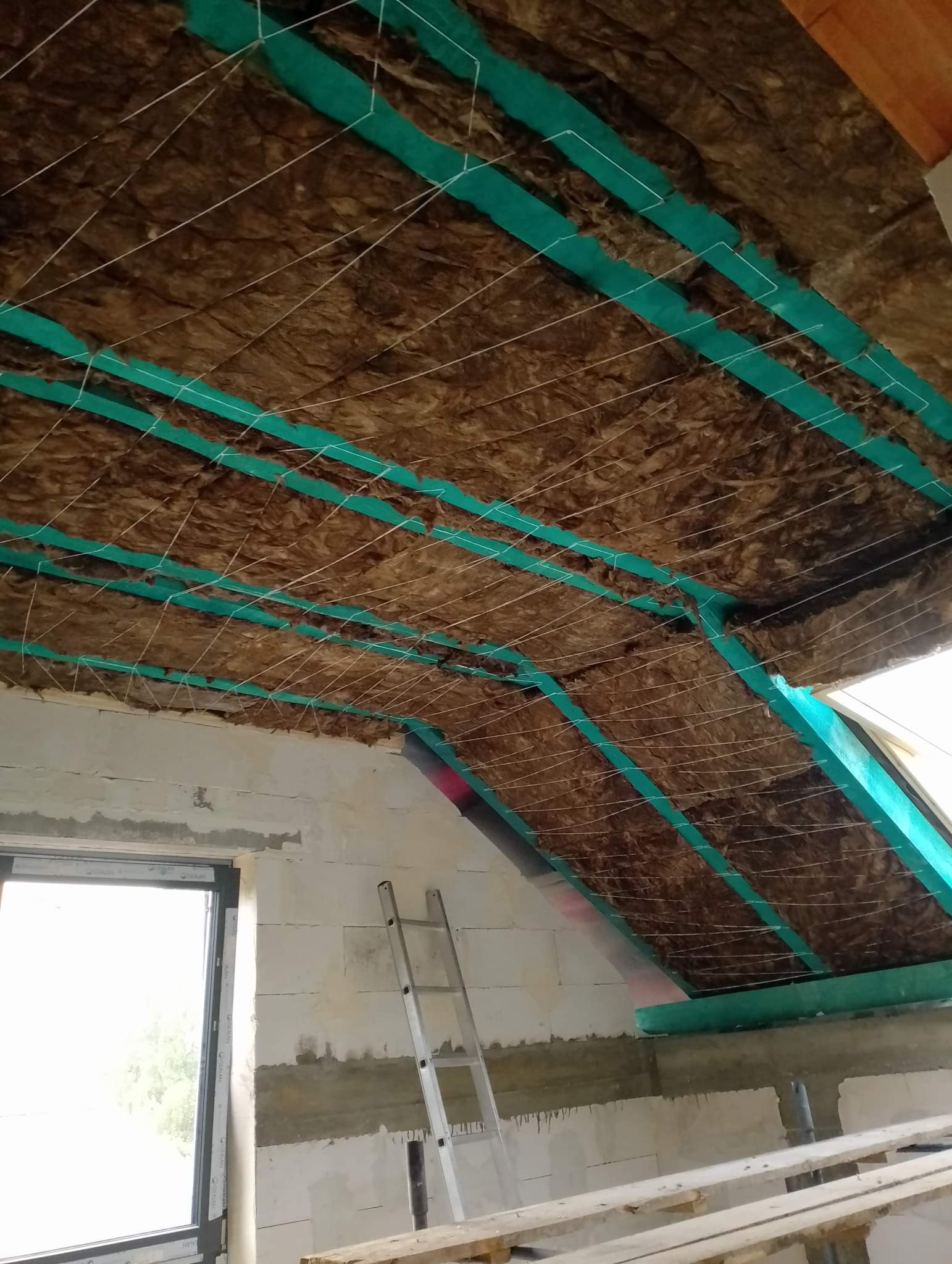 Ocieplenie poddasza wełną mineralną, zabezpieczoną sznurkiem i zielonymi taśmami, widok na surową ścianę z oknem i drabinę opartą o ścianę.