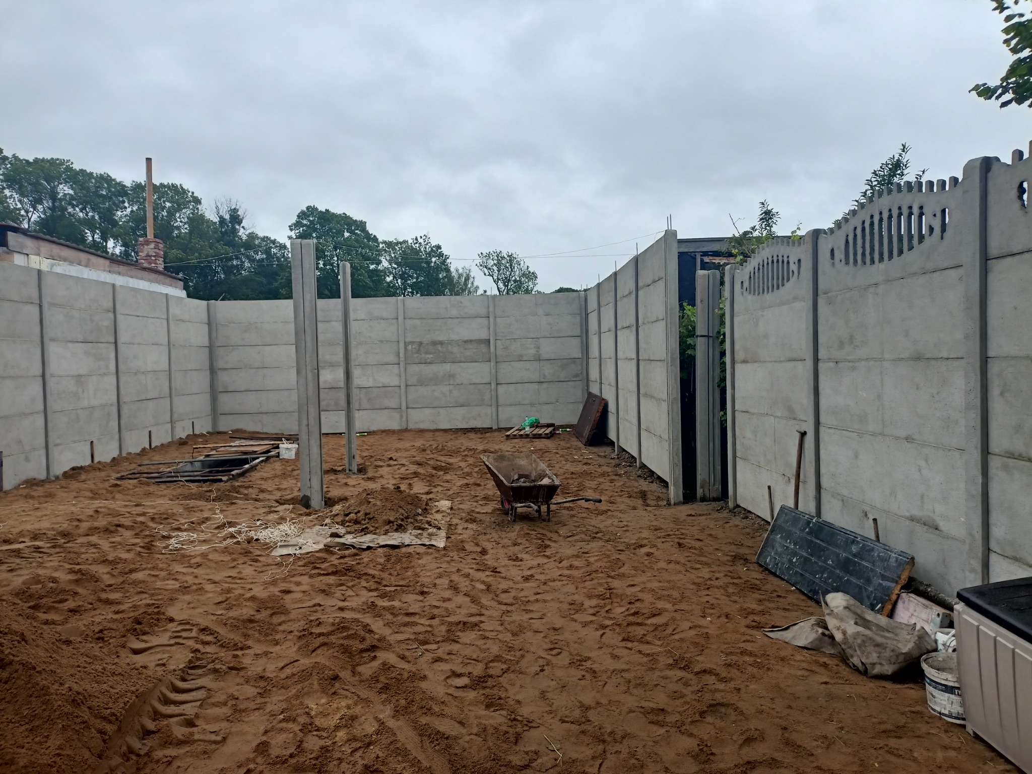 Montaż prefabrykowanego ogrodzenia betonowego, widoczne słupy i panele w trakcie instalacji, ziemia rozkopana, taczka w tle.